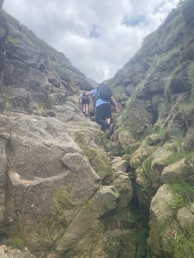 The image shows two people climbing up a steep, rocky path. The path is narrow and winding, with large boulders on either side. The people are wearing casual clothing, and one is wearing a backpack. The sky is cloudy, and the environment is a mountainous, wild area. The location is a rocky path in the Peak District National Park in Edale, United Kingdom.