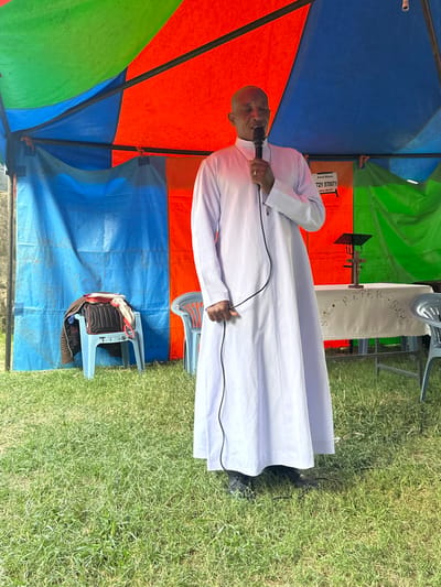 The image depicts an outdoor event taking place in Kiganjo, Kenya, under a large, multi-paneled tent with sections of blue, orange, and green fabric. The central figure is a bald man, dressed in a long, white robe or cassock, standing and speaking into a wired microphone. He holds the microphone to his mouth with his right hand and manages the cable with his left.

The event appears to be held on a sunny day, as indicated by the bright, clear lighting and the vibrant green grass that forms the ground. Behind the speaker, a table covered with a white cloth is visible, featuring the embroidered or printed text "PETER S.G.C", suggesting a religious organization like "St. Peter's Gospel Centre" or similar. Further in the background, an orange panel or banner displays the text "Band Member" and a phone number, "721 570971". To the left of the speaker, a light blue plastic chair holds some personal belongings. The scene conveys a sense of an outdoor community gathering, likely a religious service, sermon, or public address, with the speaker engaging an unseen audience.