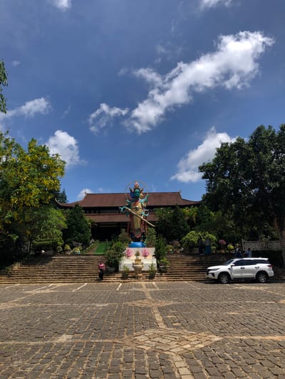 This is a wide-angle, eye-level, outdoor shot taken on a sunny day in Nam Ban, Vietnam. The image is oriented vertically. In the foreground, a large, paved area made of dark cobblestones stretches diagonally across the frame. A white SUV is parked on the left side of this paved area, which appears to be a driveway or a plaza leading to a building.
In the middle ground, a grand stone staircase ascends towards a traditional building with a brown, tiled roof. The staircase is flanked by lush greenery and decorative elements, including a statue of a colorful deity with multiple arms, adorned with gold and blue. Pink flowers are visible near the statue. Several people are scattered throughout the scene, some walking up the stairs, others near the vehicle, and a few are visible amidst the foliage.
The background features a vibrant blue sky with scattered white clouds. Large trees with dark green foliage frame the scene on the left and top, with a particularly prominent tree with yellow blossoms to the left of the building. The overall atmosphere is bright and serene, suggesting a pleasant day for sightseeing or visiting a religious site. No visible text is present in the image.