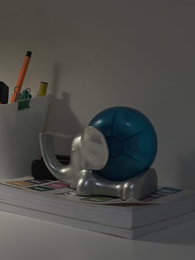 This is a close-up, low-angle, still life photograph of office supplies and a decorative object on a white surface. The scene is dimly lit, with light coming from the left, casting shadows to the right.
In the background, a light-colored wall is visible, with a subtle shadow line running horizontally.
In the mid-ground, a white cylindrical cup holds several writing implements. An orange marker with a black tip is prominently positioned, and a yellow marker or pen is also present. A green binder clip is attached to the side of the cup. Behind the cup, a dark object, possibly a phone or another office supply, is partially visible.
Dominating the foreground and mid-ground is a silver-colored decorative object shaped like an elephant. The elephant has a trunk that curves upwards and appears to be holding a translucent blue orb. The orb has a segmented design, with radial lines emanating from a central point. The elephant is resting on top of two books.
The books are stacked horizontally, with the top book having a cover with colorful squares containing stylized human figures and Hebrew text. The edges of the books are visible and appear to be white paper.
The overall impression is one of a desk or shelf with personal and office items. The image does not contain any elements that would allow for identification of a specific location within Kfar Saba, Israel.