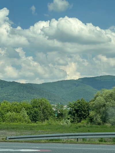 The image captures a vibrant daytime landscape in Ustroń, Poland, viewed from what appears to be a moving vehicle or roadside. The scene is dominated by a majestic mountain range covered in dense green forest, stretching across the mid-ground. Above the mountains, the sky is partly cloudy, featuring large, fluffy white cumulus clouds against patches of clear blue.
In the foreground on the right, a section of a paved road is visible, bordered by a gray metal guardrail. Beyond the guardrail, there's a grassy verge and various trees and bushes with lush green foliage. In the mid-distance, nestled among the trees on the mountainside, a cluster of white buildings or structures can be faintly discerned, suggesting a small settlement or village.
The weather appears fair and bright, indicative of late morning or early afternoon. The overall impression is one of natural beauty and tranquility, characteristic of a mountainous region. A notable detail on the road surface is a partial red and white circular road marking, which appears to be a speed limit sign, with the number "0" clearly visible within the circle, likely part of "50" or "70". No people are visible in the image.