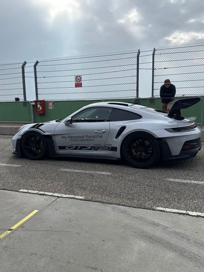 A silver Porsche 911 GT3 RS, adorned with "My Personal Porsche Track Experience" and "GT3 RS" decals, is stationary on an asphalt surface. The vehicle is positioned facing left, exhibiting black multi-spoke wheels with visible yellow brake calipers and a prominent rear wing. To the right of the car, a male individual in a dark jacket, light trousers, and sunglasses leans against a low green concrete barrier. Behind this barrier, a tall chain-link fence extends across the background. A red fire extinguisher cabinet is mounted on the green barrier in the midground, and a white circular "No Smoking" sign ("VIETATO FUMARE" in Italian) is affixed to the chain-link fence. The foreground features an asphalt section with a white demarcation line, leading to a concrete area with a yellow line. The sky above is overcast. This scene is located at a racetrack within Cervesina, Italy, likely the Tazio Nuvolari Circuit.