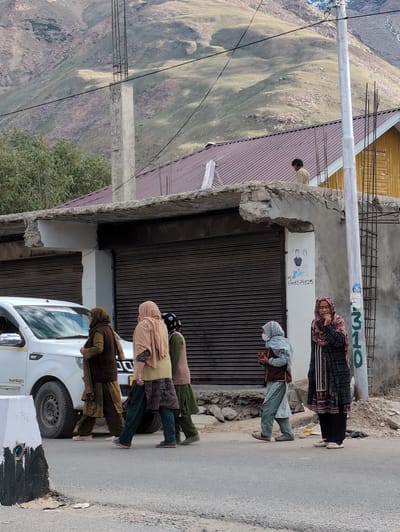 A group of five women are walking on a paved road in front of buildings. A white SUV is parked on the left side of the image. The buildings are made of concrete and have metal roll-up doors. In the background, there are mountains and trees. One woman is wearing a black and pink headscarf, a patterned long coat, and black pants. Another woman is wearing a light blue headscarf and a grey patterned outfit. A third woman is wearing a beige headscarf and a dark brown vest over a patterned tunic. A fourth woman is wearing a patterned headscarf and a patterned tunic with dark pants. The fifth woman is wearing a brown headscarf and a dark brown vest over a patterned tunic. A man is standing on top of a building in the background. The location is identified as Padum, India, based on visual cues and potentially external information.
