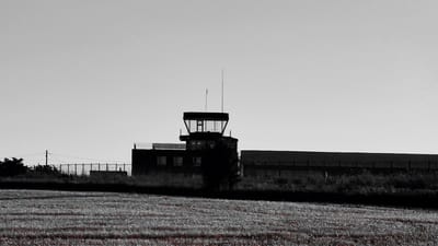 This image depicts an air traffic control tower and adjacent building silhouetted against a pale sky. The scene is set in a rural or semi-rural area, with a field of what appears to be harvested crops in the foreground. The foreground is characterized by rows of stubble, with some reddish-brown elements hinting at the previous crop.
Behind the field, a low fence with regularly spaced posts runs horizontally across the frame, separating it from a darker strip of land covered in vegetation. Beyond this, the main subject is a dark, angular building with a control tower perched on top. The control tower has a distinctive glass-enclosed cab at its apex, with antennas extending upwards. The building itself appears to be long and low-slung, with a few rectangular windows visible on its side. A flat-roofed structure with a railing, possibly a balcony or an observation deck, is attached to the side of the building.
A solitary, dark tree stands in front of the building, partially obscuring its lower section and adding depth to the composition. The overall mood is somewhat stark and industrial, with the monochromatic rendering and the clear, pale sky suggesting a neutral or possibly overcast day. The time of day is difficult to determine precisely, but the soft light could indicate early morning or late afternoon. There is no visible text in the image. The location is Horesti, Moldova.