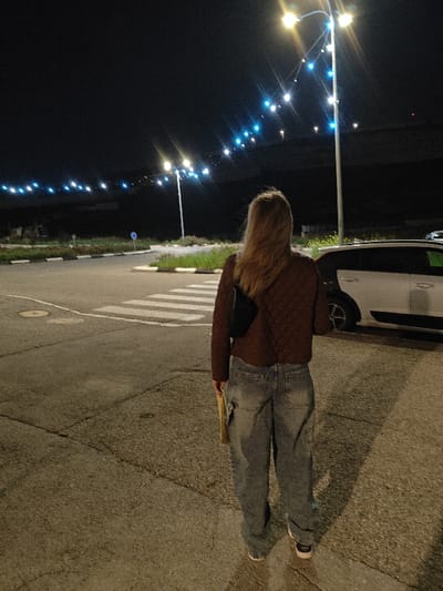 At night, a woman stands with her back to the viewer. She wears a brown, quilted jacket and baggy, light-wash jeans. A black purse hangs over her shoulder. The scene is illuminated by streetlights, which cast a warm glow on the pavement and a zebra crossing. A white car is parked to the right. In the distance, more lights line the horizon. The overall setting is urban and quiet.