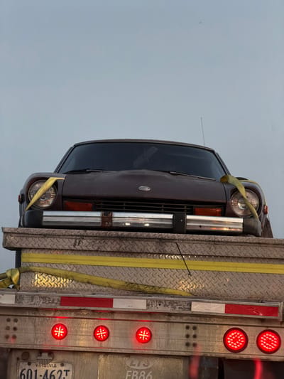 A brown car is secured to a truck bed with yellow straps. The car faces forward, its headlights visible. The truck bed is diamond-plated and has multiple red brake lights. A Virginia license plate reading "601-462T" is visible on the truck. The background is a cloudy sky.