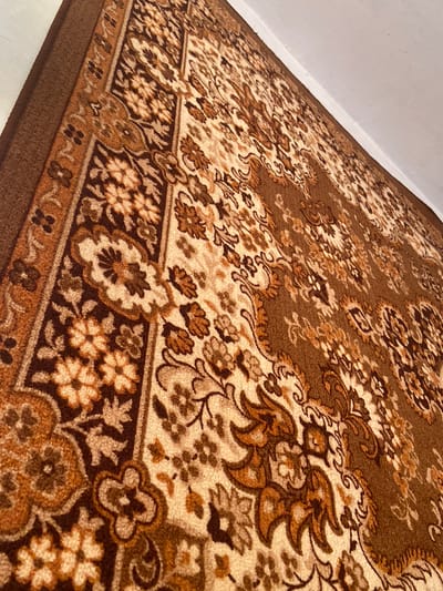 An overhead, close-up shot showcases a vintage, brown and beige floral patterned carpet. The carpet is laid on a white floor, with the corner of a white wall visible at the top right of the image. The perspective is angled, looking down and across the carpet. The intricate design features repeating motifs of flowers and scrolling vines in varying shades of brown and cream. The texture of the carpet appears plush and somewhat worn, typical of older home furnishings. The scene is static and appears to be an interior shot, likely within a residential setting in Soroca, Moldova. There are no people, actions, or interactions depicted.