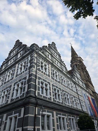This is a low-angle, eye-level shot of the Ansbach Town Hall (Rathaus Ansbach) in Ansbach, Germany, taken on a partly cloudy day. The focus is on the ornate facade of the historic building, characterized by its dark grey plaster with a distinctive white geometric pattern, resembling half-timbering. Multiple rows of windows with white frames punctuate the facade. To the right, a tall, Gothic-style tower, likely part of a church, rises against the sky. German and European Union flags hang on the side of the building near the tower. Green foliage from a tree is visible in the upper right corner of the image, and a street lamp is mounted on the building's facade. A street sign is partially visible at the bottom left, reading "Montgelaspla".