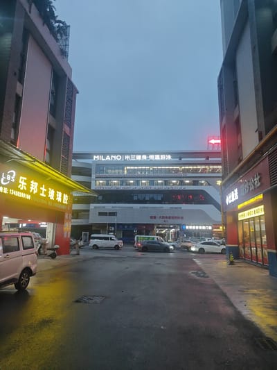 This image depicts a street scene in the Nanhai District of China, likely at dusk or early evening, given the artificial lighting and overcast sky. The foreground is dominated by a street wet from recent rain, with reflections of neon signs on the asphalt. On the left, a yellow illuminated sign advertises "乐邦士玻璃胶" (Lebangshi Glass Glue) along with a phone number and address. Next to it, a white van is parked, and further down the street, other vehicles, including a white van and several cars, are visible, suggesting a commercial or industrial area.
In the background, a multi-story building with illuminated signage stands prominently. The main sign reads "MILANO | 米兰健身·恒温游泳" (MILANO | Milan Fitness · Constant Temperature Swimming Pool), with additional text in English and Arabic below, identifying it as a "Dazhuanwan Home Furnishings & Building Materials Cross-border E-commerce Sourcing Hub" and a "center for cross-border e-commerce sourcing of building materials and furniture." A red arrow points towards the building from the right side of the frame.
The overall atmosphere is somewhat utilitarian and commercial, with a sense of ongoing activity suggested by the lighting and vehicles. The wet streets and cloudy sky create a slightly muted, possibly damp, environment. Visible text also includes names of shops like "钜隆·大转弯装饰材料城" (Julong · Dazhuanwan Decorative Materials City) and "创球盛世玻璃门窗" (Chuangqiu Shengshi Glass Doors and Windows), indicating a concentration of building and home furnishing businesses. Another sign on the right displays "神社通路 用炒公社" (Shenshe Tonglu Yongchao Gongshe), which appears to be a restaurant or food establishment, advertising "fresh ingredients, small pot stir-fry, see it to believe it."