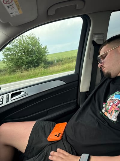 The image displays a male individual sleeping in a vehicle's passenger seat while traveling through Gabarnitsa, Bulgaria. The man has a short goatee and is wearing gold-framed sunglasses, a black t-shirt with a colorful architectural graphic on the chest, black shorts, and a dark-colored smartwatch on his left wrist. An orange smartphone, encased in a matching orange case featuring a circular design and cutouts for three camera lenses, rests on his lap. The vehicle's interior features a black door panel with stitching, a silver trim piece, a silver door handle, and buttons for window and lock controls. A grey sun visor displaying an "AIRBAG" warning label is visible above the window. Outside the window, a blurred rural landscape, characteristic of the environment in Gabarnitsa, Bulgaria, shows green trees, fields, and a section of a road under an overcast sky, indicating the vehicle is in motion.