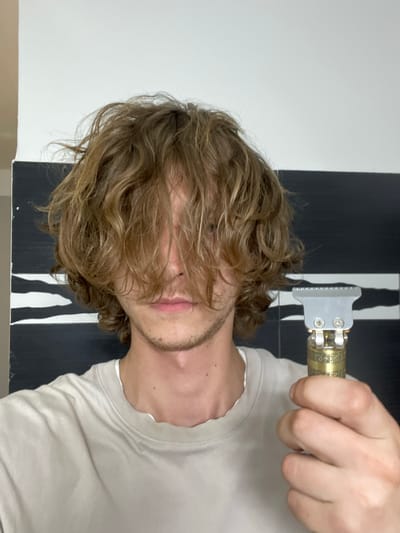 The media file is a close-up, horizontally oriented photograph of a person holding a hair trimmer. The person, who appears to be male with wavy, light brown hair partially obscuring their face, is wearing a light-colored t-shirt. The focus is on the person's hand holding a silver and gold hair trimmer, with its blades positioned near their hair. The background features a stark contrast between a black surface with white vertical stripes and a white wall. The lighting suggests an indoor setting, possibly a bathroom or a room with minimalist decor. The image composition and the close-up view of the hair trimmer imply an activity related to personal grooming, specifically hair trimming or styling. There is no visible text in the image. The image is cropped tightly on the subject and the object, offering little context about the surrounding environment beyond the black and white graphic element. The skin tone and hair color are consistent with individuals of European descent. The overall impression is one of a candid moment captured during a personal grooming routine.