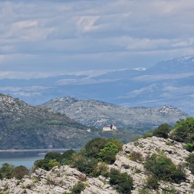 The scene is an angled outdoor shot of Godinje, Montenegro. It depicts a mountainous landscape under a partly cloudy sky. In the mid-ground, there's a mountain range with a building having a red roof. The range is sparsely vegetated. To the right, a closer mountain face shows a craggy, rocky terrain with scattered trees. A body of water is visible at the bottom of the mountain.