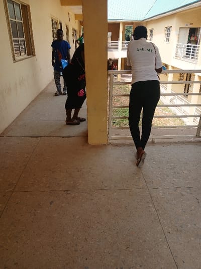 The image shows an outdoor, covered corridor or balcony on an upper floor of a multi-story building in Ugi Majowel, Nigeria. The floor is composed of light brown terrazzo with visible dark grey expansion joints.
In the foreground, a person is standing with their back to the viewer, wearing a white short-sleeved shirt with "ASK ME" printed in green on the back, black trousers, and light-colored open-toed sandals. This individual is positioned beside a cream-colored structural pillar, looking out over a white metal railing that features horizontal bars.
To the mid-left, partially obscured by the same cream-colored pillar, a second individual is visible. This person wears a dark-colored garment with pink floral patterns and light-colored open-toed sandals. Their head is turned away or out of the frame.
Further down the corridor, in the background left, a third individual is present, wearing a blue short-sleeved top and patterned black and white trousers, carrying a light blue bag on their left shoulder. This person faces generally towards the foreground.
The corridor wall on the left is light cream-colored, featuring multiple rectangular windows with white frames and horizontal security bars. Beyond the railing, across an open courtyard, another section of the multi-story building is visible. This section also features light cream-colored walls, green-tiled roofs, white balcony railings, and numerous windows and doors. Green foliage is visible in the open space between the building sections. The scene is illuminated by natural daylight.