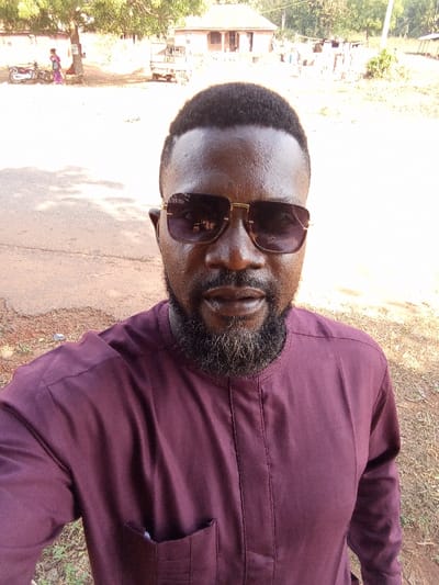 A male individual, facing forward, dominates the foreground in a selfie-style shot within Ugwolawo, Nigeria. He wears a maroon or deep burgundy long-sleeved top with a mandarin collar and a left chest pocket. His short, dark hair features a fade haircut. He has a full, dark beard and wears gold-framed sunglasses with dark lenses. His expression is neutral.

The background reveals an unpaved, reddish-brown dirt ground with sparse dry grass. In the mid-ground, a light-brown single-story building with a veranda and pillars is visible. To its left, a tree stands, beneath which a white motorcycle is parked. Two individuals, one appearing to be a woman in a purple and white patterned dress, are standing near the motorcycle. Further to the right of the main building, stacked cement blocks are present, along with indistinct structures and additional vegetation. A light-colored small truck or cargo tricycle is partially visible between the tree and the main building. The scene is illuminated by bright daylight, casting shadows in the foreground.