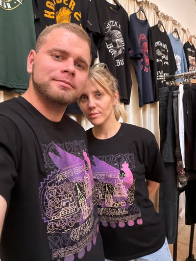 In a dimly lit room, two individuals, a man and a woman, pose for a selfie, both wearing identical black t-shirts with a purple and gold graphic. The graphic on the t-shirts depicts a stylized representation of the city of Suzdal, Russia, featuring prominent landmarks such as the Suzdal Kremlin and the bell tower of the Church of the Nativity of the Blessed Virgin Mary. The text on the shirts appears to be in Russian and mentions "Всероссийский турнир" (All-Russian Tournament) and "Финал Чемпионата" (Championship Final). Behind them, a rack of t-shirts and denim pants are neatly arranged, suggesting a retail setting or a personal collection. The background is dominated by a beige curtain. The man, with light brown hair and a beard, has his arm around the woman, who has blonde hair and is wearing a necklace. Their expressions are relaxed and engaging.