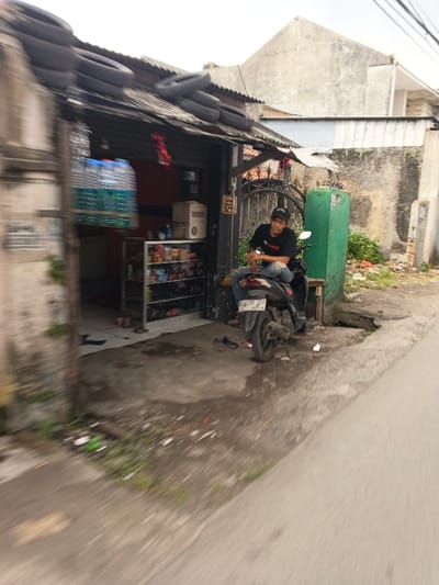The image depicts a roadside scene in Margahayu, Indonesia. A male individual, wearing a black t-shirt, ripped blue jeans, and a black baseball cap, is seated sideways on a dark-colored scooter or motorcycle, license plate D 6735 ZCY, parked on a dirt and concrete patch. He is looking to his left, away from the camera. To his left is a small, open-fronted shop with a corrugated metal roof, on which several black tires are stacked. Inside the shop, a metal shelving unit displays various packaged goods, and multiple large water bottles are visible near the entrance. A light-colored appliance is also present within the shop. To the right of the shop and behind the man is a dark metal gate, followed by a green concrete pillar. The ground in the immediate foreground consists of asphalt road transitioning to a dirt and cracked concrete area with scattered litter and sparse vegetation. In the background, a weathered concrete wall separates the area from an overgrown patch, and a light-colored residential building with a distinct peaked roof is visible under an overcast sky. Overhead power lines are faintly discernible. The image exhibits motion blur, indicating it was captured from a moving perspective.