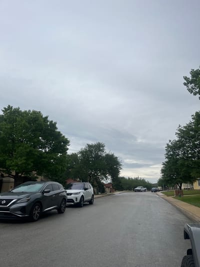 The image depicts a residential street in San Antonio, United States of America. Two cars are parked along the right side of the street, a gray Nissan Murano and a white Land Rover Range Rover. Other vehicles are visible further down the street. The sky is overcast. The road appears wet. Trees line both sides of the street. Houses are visible behind the trees.