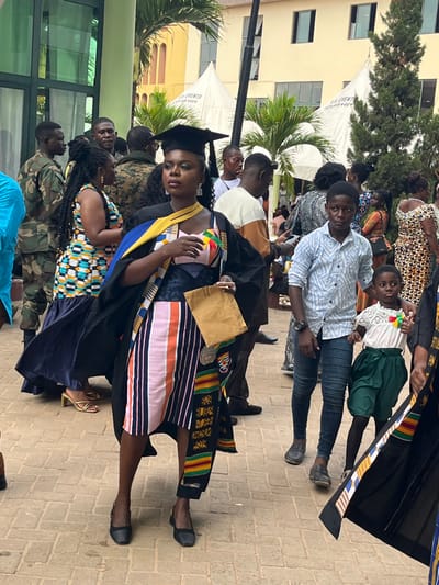 An outdoor scene depicts a crowded event, likely a graduation ceremony, in Madina, Ghana. The central figure is a young Black woman, approximately mid-20s, standing upright and looking slightly to her left. She wears a black academic gown over a strapless top featuring geometric patterns in yellow, blue, red, and white, paired with a vertically striped skirt in pink, orange, white, and navy. A yellow and blue stole is draped around her neck, and a Kente cloth sash with black, red, yellow, and green patterns hangs from her left shoulder. She wears a black academic cap and black low-heeled shoes. Her right hand holds a light brown envelope.
To her immediate right, a young Black boy, approximately 10-12 years old, wears a light blue patterned long-sleeved shirt and dark jeans, with his right hand placed on the shoulder of a smaller Black child, likely a girl, aged around 4-6. The smaller child wears a white lace-trimmed top with a small accessory displaying Ghanaian flag colors (red, yellow, green, black) and green pleated shorts/skirt.
The background shows numerous individuals of varying ages and attire, including men in military camouflage uniforms, a woman in a colorful patterned dress with gold-colored high heels, and other attendees. Several white event tents labeled "M & M EVENTS" are visible behind the crowd. Modern buildings with light green and beige facades, large windows, palm trees, and evergreen trees form the distant background. The ground is paved with rectangular light-colored bricks. The lighting suggests a bright daytime setting.