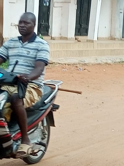A man is riding a motorcycle in Wuro Dole, Nigeria. He is wearing a striped shirt and shorts. The motorcycle is red and black. The background is a sandy road with some buildings.