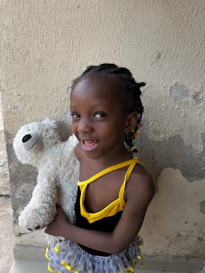 A young, dark-skinned girl in a black and yellow top and gray tutu is holding a white stuffed polar bear. She is smiling and looking up at the camera. Her hair is in braids decorated with colorful beads. The girl is sitting in front of a wall with peeling paint. The location is Gauraka, Nigeria.