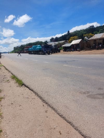 The image displays a daytime scene in Kimamba, Tanzania, featuring a two-lane asphalt road with a dirt shoulder on its left side. A blue semi-truck loaded with teal-colored bundled cargo is parked on the right side of the road, followed by another lighter-colored truck. Further along the right, a row of single-story structures with various roof types (corrugated metal, possibly tile) lines the road, fronting a densely vegetated hillside. Several individuals are visible near these buildings, some standing and some seated. A lone pedestrian, clad in dark clothing, is captured mid-stride, walking from left to right across the asphalt road in the middle ground. A portion of a bicycle is visible on the far left edge of the dirt shoulder. The sky above is clear blue with scattered white cumulus clouds.