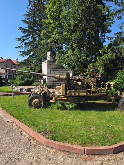 The image depicts an anti-aircraft gun displayed on a grassy area alongside a paved road. The gun is a light khaki-green color, appearing to be a model from the World War II era. In the background, a monument is visible, partially obscured by a tree; the monument seems to be a bust atop a stone base with inscribed text. Beyond the gun and monument, there is a glimpse of a building with a red-tiled roof, indicating an urban setting. The sky is clear blue. This location is in Barziya, Bulgaria.