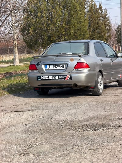A silver four-door sedan, identified as a Mitsubishi by its rear emblem and "MITSUBISHI SPORT" decal on the bumper, is parked on an uneven, broken asphalt and gravel surface. The vehicle bears a Bulgarian license plate, "A 1929 HT." It features a small rear spoiler, a "rock on" skeleton hand sticker on the left side of the trunk lid, and a Harley-Davidson logo sticker on the right. White pinstriping runs along the side body, and a "SPORT" decal is present on the lower rear side panel. A sunshade is visible through the rear window. The vehicle's exhaust pipe protrudes from the right rear. No individuals are visible.
The scene is an outdoor environment in what is identified as Kermen, Bulgaria, based on the license plate's regional indicator 'A' for Sliven, which includes Kermen. The ground consists of a degraded road surface with visible cracks, potholes, and loose gravel. To the left of the vehicle, a grassy verge transitions into bare deciduous trees, and a stone wall is partially visible further back. In the background, behind the car, are several tall, green coniferous trees and what appears to be a utility pole or similar structure on the far right. The lighting suggests direct sunlight, casting shadows.