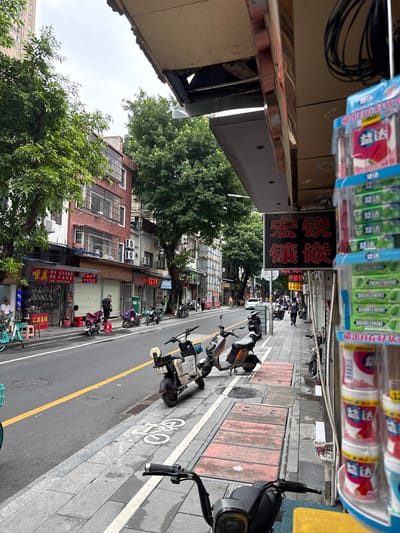 A street scene in Liwan District, China, shows a sidewalk lined with shops and parked motorcycles. In the foreground, a black and white electric motorcycle with license plate A65082 is parked facing left. The sidewalk has a bicycle lane marked with white paint. To the right, a display rack holds various products, including packages of mints and other small items. A red sign with Chinese characters is mounted on the building above the sidewalk. Several shops have open storefronts, displaying goods and signs. A man in a black t-shirt and dark pants walks away from the camera on the sidewalk, past a covered red electric scooter. Further down the street, more motorcycles and scooters are parked. Trees line the street, providing shade. Buildings of various heights and architectural styles are visible, some with exposed wires and air conditioning units. The sky is overcast.