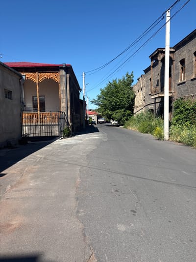 The image depicts a street scene in the city of Ashtarak, Armenia, under a clear blue sky. The street is paved with asphalt, and the sidewalk on the left is also asphalt. On the left side of the street, there is a building with a red roof and a detailed wooden balcony. Next to it is a simpler, gray building with a window. A black metal fence encloses a small area in front of the building with the red roof.

On the right side of the street, there is an older, dark stone building with balconies and intricate wooden details, suggesting historical architecture. Greenery, including tall grass and bushes, grows along the side of this building. A white utility pole with electrical wires stands in front of it. In the distance, further down the street, more buildings and trees are visible, and a white vehicle and a light-colored truck are parked or moving. The scene appears to be a residential or older commercial area in Ashtarak.