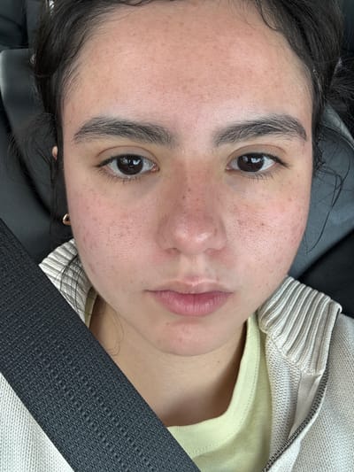 A close-up photograph depicts a young woman with dark hair, light skin, and brown eyes. Her face is visible, showing freckles and natural features. She appears to be wearing light makeup. She has on a yellow t-shirt, with a zip-up hoodie on top. A black seatbelt is across her chest, suggesting she is in a vehicle. The background is dark and out of focus. There is no verifiable information to place the photograph in Elgin, United States of America.