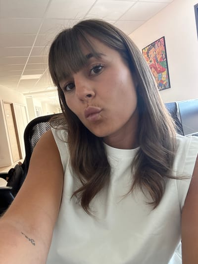 A woman is sitting in a black office chair in an indoor environment, possibly an office. She is facing the camera with a slightly tilted head, pouting her lips. She has brown hair with bangs and is wearing a white top. On her left arm, there is a small, dark tattoo. On the wall behind her, there is a colorful framed picture. Based on the location provided (Les Arcs, France), the presence of office furniture, and general indoor aesthetic, the location is likely an office interior in Les Arcs, France.