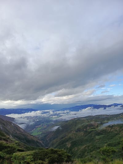 The image shows a mountainous landscape with a valley below, likely located in the vicinity of El Cisne, Ecuador. The sky is overcast with heavy, grey clouds dominating the upper portion of the image. Distant mountains are visible, partially obscured by a layer of clouds or fog. The valley contains patches of green fields and a river or stream winding through it. Patches of fog are visible in the valley and on the mountain slopes. The foreground features a steep hillside covered in dense vegetation. Some smoke or mist is visible on a hillside.