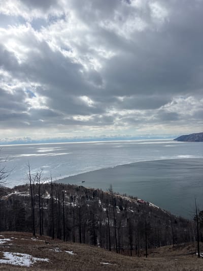 The image depicts a panoramic, elevated view of Lake Baikal in Nikola, Russia, under an overcast sky. The upper half of the frame is dominated by dense, grey cumulus clouds with intermittent brighter patches.
The middle ground features Lake Baikal, distinctly bisected by a partial ice cover. The left side of the lake displays broken, partially frozen ice reflecting light, while the right side is open, dark grey-green water with visible ripples. A clear demarcation exists between the ice and open water. In the distance, a range of snow-capped mountains stretches along the horizon beyond the lake. Two dark-colored boats are present on the open water; one larger vessel moves towards the right, leaving a wake, and a smaller object rests near the ice edge. Several unidentifiable small, dark specks are visible on the ice surface.
The foreground and mid-ground consist of a steep, undulating hillside descending to the lake. The immediate foreground is covered with sparse, dry brown grass and patches of melting snow. Numerous dark, bare trees, some appearing fire-damaged, rise from this terrain. Further down the hillside, the forest becomes denser, comprising primarily bare trees with some evergreen foliage. Along the lake's shoreline, at the base of the hill, a complex of light-colored buildings is visible on the left, and a cluster of buildings with distinct red roofs is situated on the right. A road or path parallels the water's edge. No individuals are discernible within the scene.