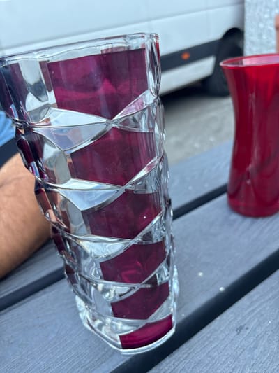 An outdoor, close-up, eye-level shot features two vases on a grey, slatted wooden table. The vase in the foreground is a tall, clear glass vase with a geometric, faceted design, revealing deep purple inserts within its structure. The vase on the right is a shorter, red glass vase with a curved silhouette. In the background, a white vehicle is partially visible. The scene appears to be set in the city of Boom, Belgium, specifically within the vicinity of a park or outdoor gathering area, suggested by the presence of similar tables and the overall ambiance.