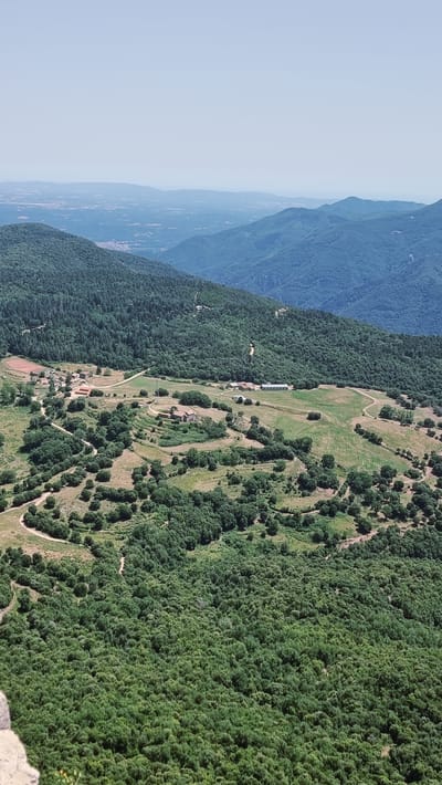 The image depicts a panoramic aerial view of a rural landscape in the municipality of Susqueda, Spain. The foreground is dominated by a dense forest of green trees. As the view recedes, the terrain opens into rolling hills with scattered trees and patches of grassland. Several buildings, appearing to be farmhouses or rural dwellings, are visible on the hillsides. A dirt road winds through the landscape, connecting the various structures. In the background, a series of progressively bluer mountain ranges stretch towards the horizon, with a hazy sky above. The overall impression is one of a tranquil, expansive natural environment with a scattered human presence.