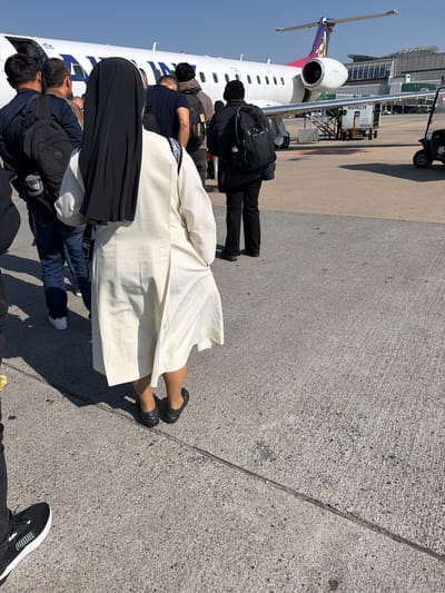 The image shows a group of people disembarking from an airplane on a sunny day at what appears to be an airport tarmac, possibly in Kempton Park, South Africa. The perspective is from behind the people, looking towards the airplane and the ground. The airplane is white with colorful branding on its tail. Several individuals are visible, some wearing backpacks, indicating they are passengers. One person in the foreground is wearing a long, flowing white garment, and another is wearing a black head covering and a black garment. The ground is paved concrete. In the background, a building with a glass facade and a jet bridge are visible. A small utility vehicle is also parked near the airplane. The scene suggests the process of deplaning and heading towards the terminal. The lighting indicates it is daytime. There is text visible on the airplane and on a piece of equipment near the airplane. The text on the airplane reads "TATA". The text on the equipment reads "MBW239" and "MB-BC02".