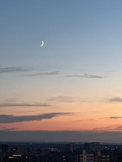 A crescent moon is visible in the upper left quadrant of the image against a clear blue sky that transitions to hues of orange and pink near the horizon. Below the sky, a city skyline is depicted at dusk. Numerous buildings of varying heights and architectural styles are present, some illuminated by interior lights. In the distance, faint industrial structures with chimneys can be observed, along with atmospheric haze. The overall scene suggests an urban environment at twilight.