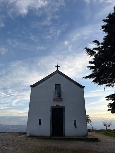 The Chapel of Senhora do Socorro, located in Peso da Régua, Portugal, is centrally framed under a partly cloudy sky. The structure is a white-painted, single-story building with a dark-colored gabled roof, topped by a black Latin cross. A dark, rectangular main entrance door is centered on the lower facade. Above the main door, a smaller, dark-framed window is present, fronted by a curved iron balcony railing. Two narrow, vertical, rectangular openings are positioned symmetrically on either side of the main entrance. The foreground consists of compacted dirt and gravel. To the right, a large, dark evergreen tree partially obscures the upper right corner. In the mid-ground, towards the right, two smaller, bare deciduous trees are visible. The background reveals a vast landscape, likely a valley or distant city, extending to the horizon under the pale blue sky marked with streaky white clouds. The lighting suggests early morning or late afternoon conditions. No individuals or specific actions are observed.