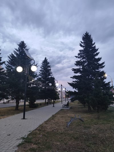 This media file depicts an outdoor scene in Slavgorod, Russia, likely during dusk or twilight. A paved walkway, constructed with grey rectangular pavers, runs diagonally from the left foreground towards the right middle ground. Flanking the walkway is a strip of dry grass interspersed with some sparse green vegetation.
Several large evergreen trees, appearing to be fir or spruce, dominate the landscape. Their dark green foliage contrasts with the muted tones of the sky. A series of lampposts are visible along the walkway. The closest lamppost features two spherical, illuminated lamps and a solar panel at its apex. A string of small, decorative lights hangs from this lamppost, adding a festive touch. Further down the walkway, other lampposts with single illuminated spheres are visible.
In the middle ground, a park bench with an ornate metal design sits on the grassy area. Beyond the trees on the left, glimpses of buildings suggest an urban or suburban setting. The sky is overcast with heavy, grey clouds, indicative of cloudy or impending rain. The overall mood is tranquil and somewhat somber due to the low light conditions and the cloudy sky. No people are visible in the frame. A sign with a circular shape and what appears to be the number "20" in white on a red background is visible on a lamppost in the far right background.