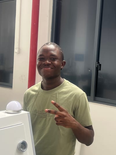 A young Black male, with braided hair and a light green t-shirt, smiles at the camera and flashes a peace sign with his right hand. He stands indoors, next to a white cabinet-like object topped with a glowing white dome light. Behind him, a large window with dark-tinted panes reflects a blurred image, and a red vertical pipe runs along a cream-colored wall. The image is a medium shot, with the subject centered and taking up most of the frame.