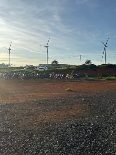 The image depicts an outdoor scene in Phát Chi, Vietnam, under a clear sky with scattered clouds. In the background, three wind turbines stand tall against the horizon. In the midground, a line of parked motorcycles is visible, suggesting a gathering or parking area. Behind the motorcycles, a dark-colored car and a white pickup truck are parked. The terrain in the foreground is a mixture of dark gravel and reddish-brown dirt. The overall environment appears to be a rural or semi-rural area with agricultural land or open fields, as indicated by the presence of greenery and the distant trees. The lighting suggests it is either early morning or late afternoon, with the sun casting a warm glow.