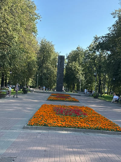 In the city of Yazvitsevo, Russia, this image captures a sunlit park scene dominated by lush green trees and a clear blue sky. In the foreground, a paved walkway transitions from grey to red brick patterns. Two rectangular flower beds are prominently featured on the walkway, the closer one filled with vibrant orange marigolds and a cluster of red flowers in its center, while a second, slightly further bed displays similar orange flowers. A tall, dark, monolithic monument stands in the background, flanked by trees. Several people are visible in the park: a couple walks on the left side of the walkway, and further back, a group sits on benches to the right, with a few individuals also walking or standing. The overall atmosphere is serene and public, suggesting a well-maintained urban park environment.