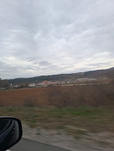 The image depicts an exterior scene likely captured from a moving vehicle in the vicinity of Cherniche, Bulgaria, under an overcast sky. In the immediate foreground, the black casing and reflective surface of a vehicle's side mirror are visible on the lower left, partially obscuring a blurred grey road and a strip of green and brown roadside vegetation. The midground features a large expanse of tilled, reddish-brown earth, characteristic of an agricultural field. Beyond this field, an industrial site or quarry is visible, characterized by multiple large, irregularly shaped piles of material, predominantly reddish-brown and light grey aggregates. Within this site, several structures, possibly buildings or machinery, are discernible. The background consists of a tree-covered hillside, rising towards a larger, distant hill or mountain range under the grey, cloudy sky. No identifiable individuals are present.
