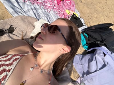 A woman is lying on a striped towel on a sandy beach. She wears sunglasses and a red and white patterned bikini top with a delicate body chain. Her head is turned to the left, and assorted items, including a blue tote bag and dark-colored clothing, are scattered nearby. Another pair of sunglasses is present near her torso. The scene suggests a relaxed day at the beach.