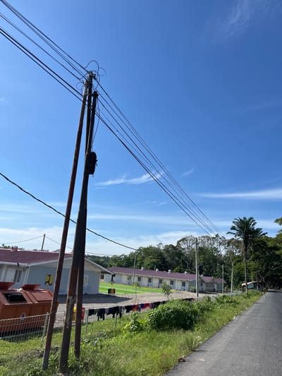 This image depicts an outdoor scene on a clear, sunny day, likely in Bukitrata, Indonesia. The primary subjects are a utility pole with multiple wires stretching across the frame and a row of low-rise buildings with red roofs in the background. The buildings appear to be part of an institution or complex, possibly a school or dormitory, given the uniformity of their design and the presence of laundry hanging on a line between poles in front of them.

In the foreground, the weathered utility pole dominates the left side of the image, with thick black wires fanning out across the bright blue sky. Wispy white clouds are scattered in the sky, indicating fair weather. To the right of the buildings, a paved road runs alongside a strip of lush green grass and vegetation. Trees, including a palm tree, line the distant background, suggesting a tropical or subtropical climate.

No people are clearly visible or identifiable in the image, though the hanging laundry implies human presence. The scene is static, with no specific activity or event taking place, rather capturing a snapshot of daily life. The overall impression is one of a peaceful, functional environment under clear skies. No discernible text is visible in the image.