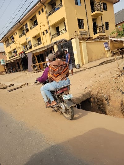 This image captures a street scene in Puerto López, Colombia, under clear daylight conditions. In the midground, a dark-colored motorcycle is depicted in motion, traversing an unpaved, sandy section of road. Two individuals are on the motorcycle: the driver, largely obscured, wears a purple shirt and a light-colored cap. Behind the driver, a female passenger wears light blue jeans and a distinctive orange, brown, and white patterned scarf or wrap over her shoulders, with bare feet resting on the motorcycle's footrests. The motorcycle displays a license plate with the alphanumeric sequence "MA 766G6".
To the immediate right of the motorcycle, a significant, deep gully or open drain with concrete edging runs parallel to the unpaved road. The foreground shows a paved road surface with visible shadows. In the background, a prominent three-story yellow building is visible, featuring multiple balconies with black railings and dark-framed windows. The building's property is secured by a concrete fence topped with barbed wire, and a black metal gate is partially open. A male individual, dressed in a blue sleeveless top and light-colored shorts, stands near this gate, observing the scene to his left. Overhead, multiple utility wires or cables are visible across the top of the frame. The environment presents a blend of paved and unpaved road infrastructure within an urban or semi-urban setting.