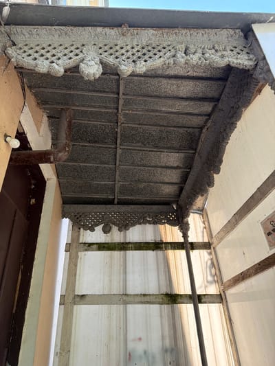 The image shows an exterior view of a porch roof with ornate decorative trim. The trim is made of a light-colored material, possibly plaster or concrete, and features a lattice pattern with carved floral motifs and rounded elements. The underside of the roof is constructed with metal panels supported by metal beams. A downspout is visible on the left side, running down the wall of the building. The building exterior appears to be stuccoed. To the right of the porch, a translucent corrugated material forms a side enclosure, supported by wooden beams. There are signs of wear and tear, including some moss or algae growth on the wooden beams and a general weathered appearance of the materials. The scene is illuminated by natural light, casting shadows and highlighting the textures of the surfaces. The location is identified as Taganrog, Russia.