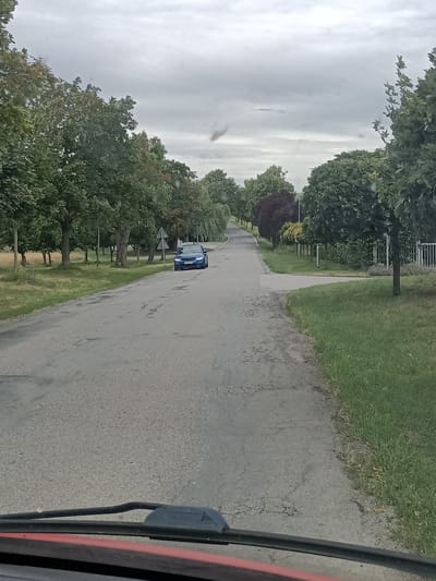 This is a photograph taken from inside a vehicle, looking out at a rural road in Svémyslice, Czechia. The scene depicts a slightly overcast day, likely in the late morning or afternoon, given the diffused natural light. The road is paved but shows signs of wear with cracks and patches.
The main subject in the scene is a bright blue car driving away from the camera on the right side of the road. Trees line both sides of the road, with lush green foliage indicating a season like summer or early autumn. On the right, there's a white fence and manicured grass, suggesting a residential or cultivated area. On the left, the landscape is more natural, with a field bordering the trees.
The activity is simply driving on a road. There are no people visible in the frame. Notable details include the overcast sky, the condition of the road, and the variety of trees, some of which are larger and more mature, while others appear to be more ornamental, like the clipped trees along the right side. The perspective from within the vehicle is evident from the dashboard and windshield wipers visible at the bottom of the frame. The overall mood is calm and mundane, depicting a typical drive through the countryside. No visible text is present in the image.