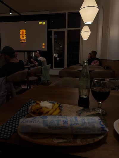 The image depicts a dimly lit indoor scene, likely a restaurant or bar, with a large screen displaying the FIFA World Cup 2026 logo and "BAR TV MUNDIAL." Several people are seated at tables, some are engaged in conversation, and others are looking towards the screen. In the foreground, a wooden table holds a meal consisting of a wrapped sandwich or burrito, a bowl of french fries with dipping sauce, a bottle of Coca-Cola, and a glass of red wine. The overall ambiance is casual and social, suggesting patrons are gathered to watch a sporting event. The location is identified as Santiago De Compostela, Spain.