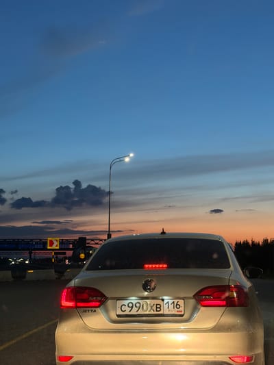 The image shows the rear of a silver Volkswagen Jetta parked on a road at dusk. The car has a Russian license plate displaying "C990XB 116". The sky is a gradient of blue and orange, with some clouds. A street light with two lamps is visible on the right side of the image. In the background, there are some traffic signs, barriers, and trees. The overall impression is a quiet roadside scene during twilight. The location is specified as Derzhavino, Russia.